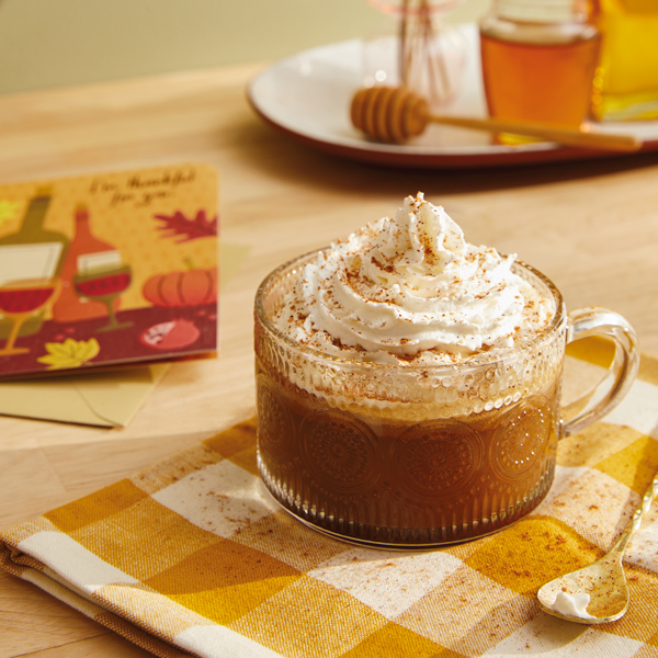 A clear glass mug with a boho pattern on the outside filled with hot buttered rum and topped with whipped cream and sprinkled with nutmeg; a thank-you card featuring a fall illustration on the front lays nearby.